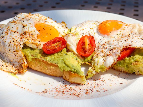 The image shows toast with avocado, cherry tomatoes, and fried eggs on top, garnished with a spice, on a white plate.