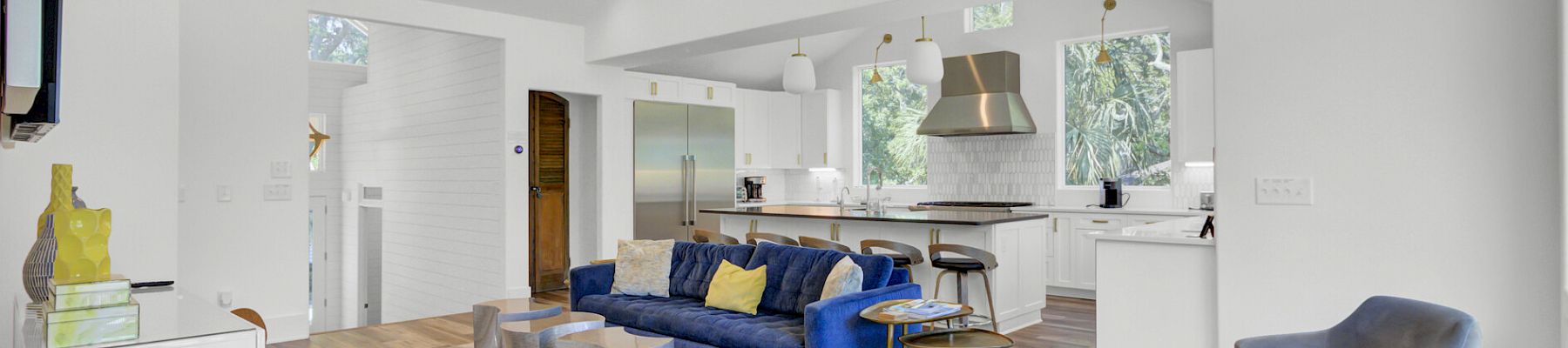 A modern living room with a blue sofa, stylish chairs, decorative lighting, and a view into a kitchen area, featuring sleek furnishings.