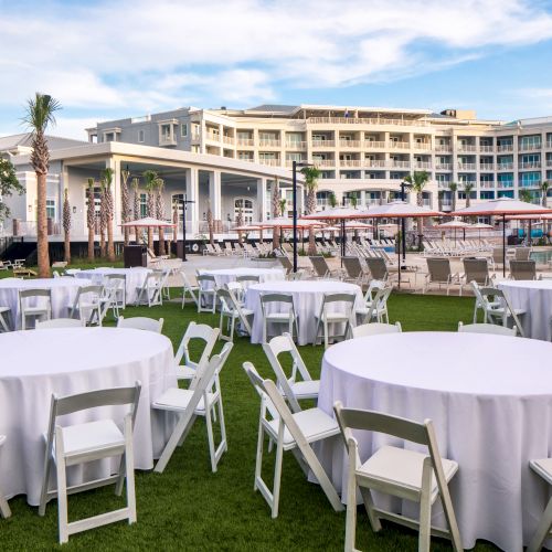 Outdoor event setup with round tables, white tablecloths, and chairs on a lawn near a resort building and pool area.