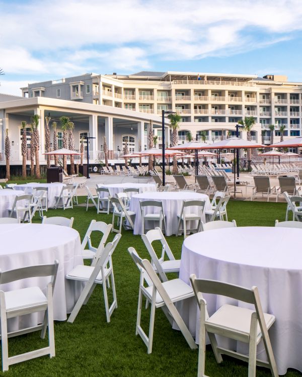 Outdoor event setup with round tables, white tablecloths, and chairs on a lawn near a resort building and pool area.