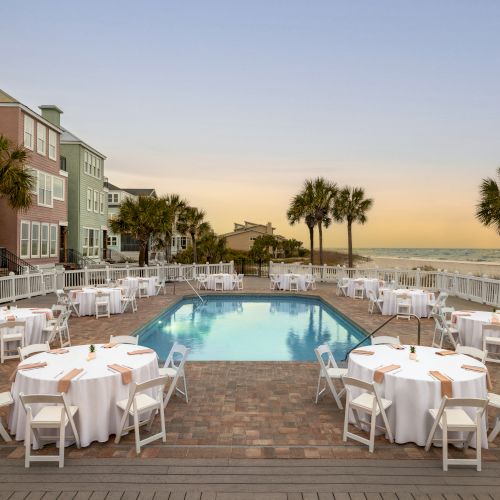 A poolside setup with round tables and white chairs, surrounded by colorful buildings and palm trees, overlooking the ocean at sunset.