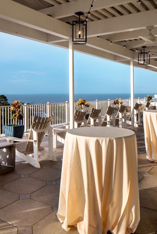 An outdoor terrace with tables covered in cloth, wooden chairs, and potted plants overlooking the sea.