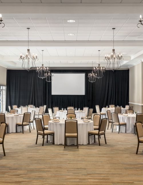 A banquet hall with round tables and chairs, set for an event with chandeliers above and a large screen at the front.