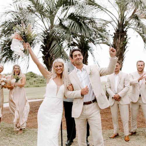 A newlywed couple celebrates joyfully outdoors, surrounded by friends and palm trees in a relaxed, tropical setting.