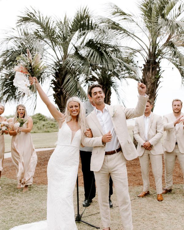 A newlywed couple celebrates joyfully outdoors, surrounded by friends and palm trees in a relaxed, tropical setting.