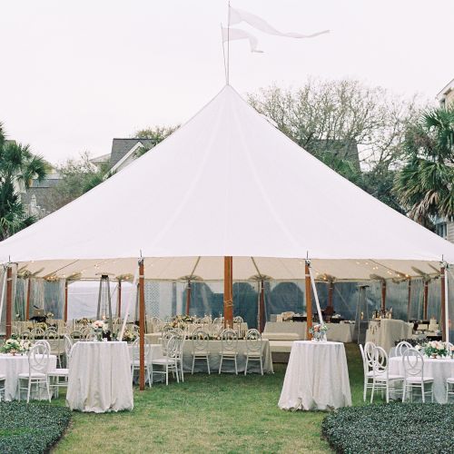 A white tent set up outdoors with tables and chairs, surrounded by palm trees and houses, likely for an event or gathering.