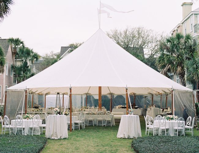 A white tent set up outdoors with tables and chairs, surrounded by palm trees and houses, likely for an event or gathering.