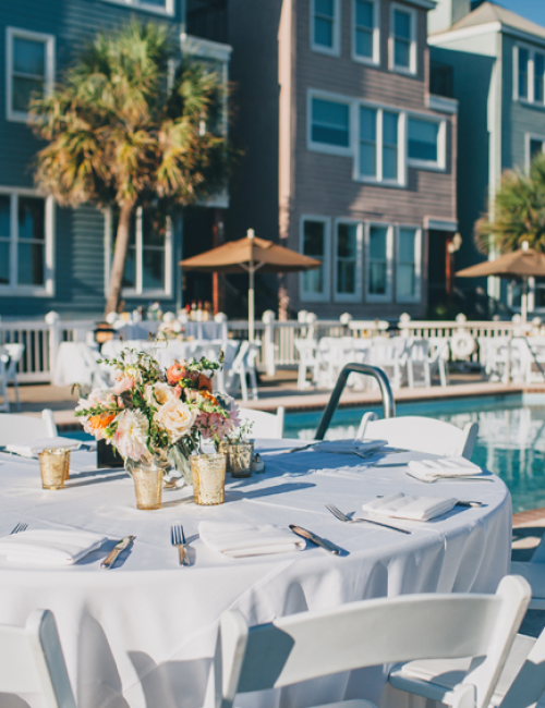 An outdoor event setup with round tables and white chairs, poolside, near colorful buildings and palm trees.