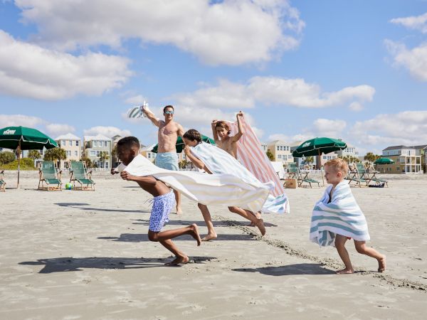 A group of people, including children with towels, play on a sunny beach, with umbrellas and houses visible in the background.