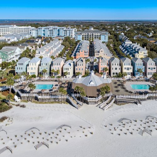 A coastal resort with colorful houses, pools, and beach umbrellas on a sandy beach, surrounded by greenery and buildings in the distance.