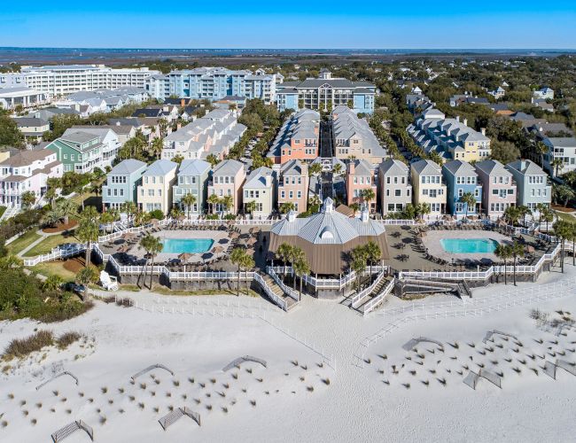 A coastal resort with colorful houses, pools, and beach umbrellas on a sandy beach, surrounded by greenery and buildings in the distance.