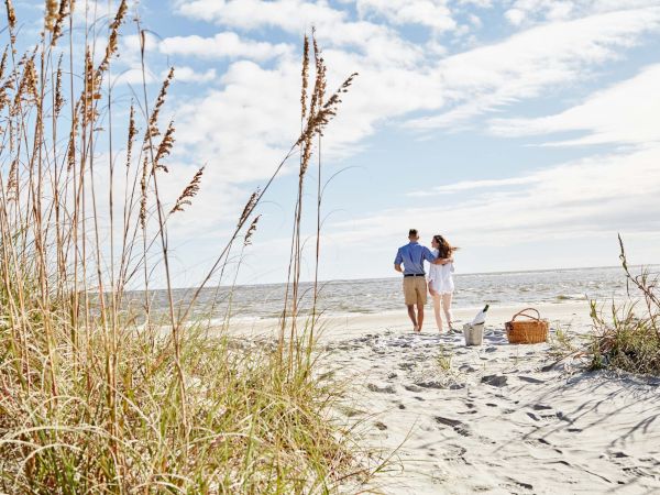 A couple is walking on the beach near the shore, with sea grass in the foreground and a picnic setup on the sand.