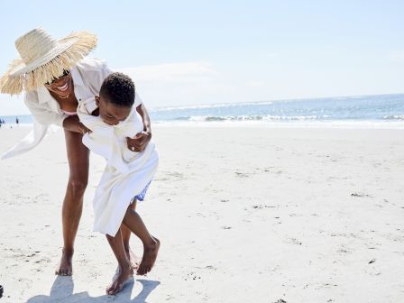 A person and a child are playing joyfully on a sunny beach, with the ocean and a clear sky in the background.