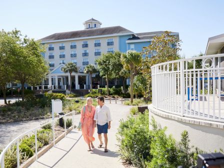 A couple walks on a path in front of a large, blue-and-white building surrounded by greenery and a clear sky.