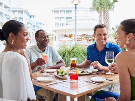 Four people sit at an outdoor restaurant table, enjoying drinks and food, while smiling and chatting with each other.