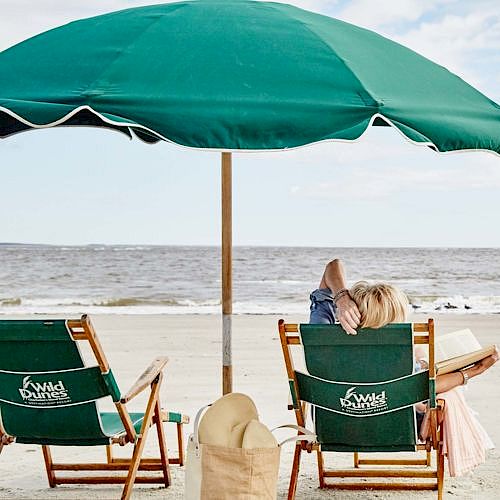 A beach scene with two chairs under a green umbrella facing the ocean. A person relaxes in one chair, next to a straw hat and a bag.