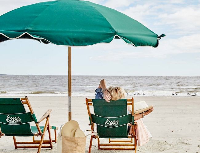 A beach scene with two chairs under a green umbrella facing the ocean. A person relaxes in one chair, next to a straw hat and a bag.