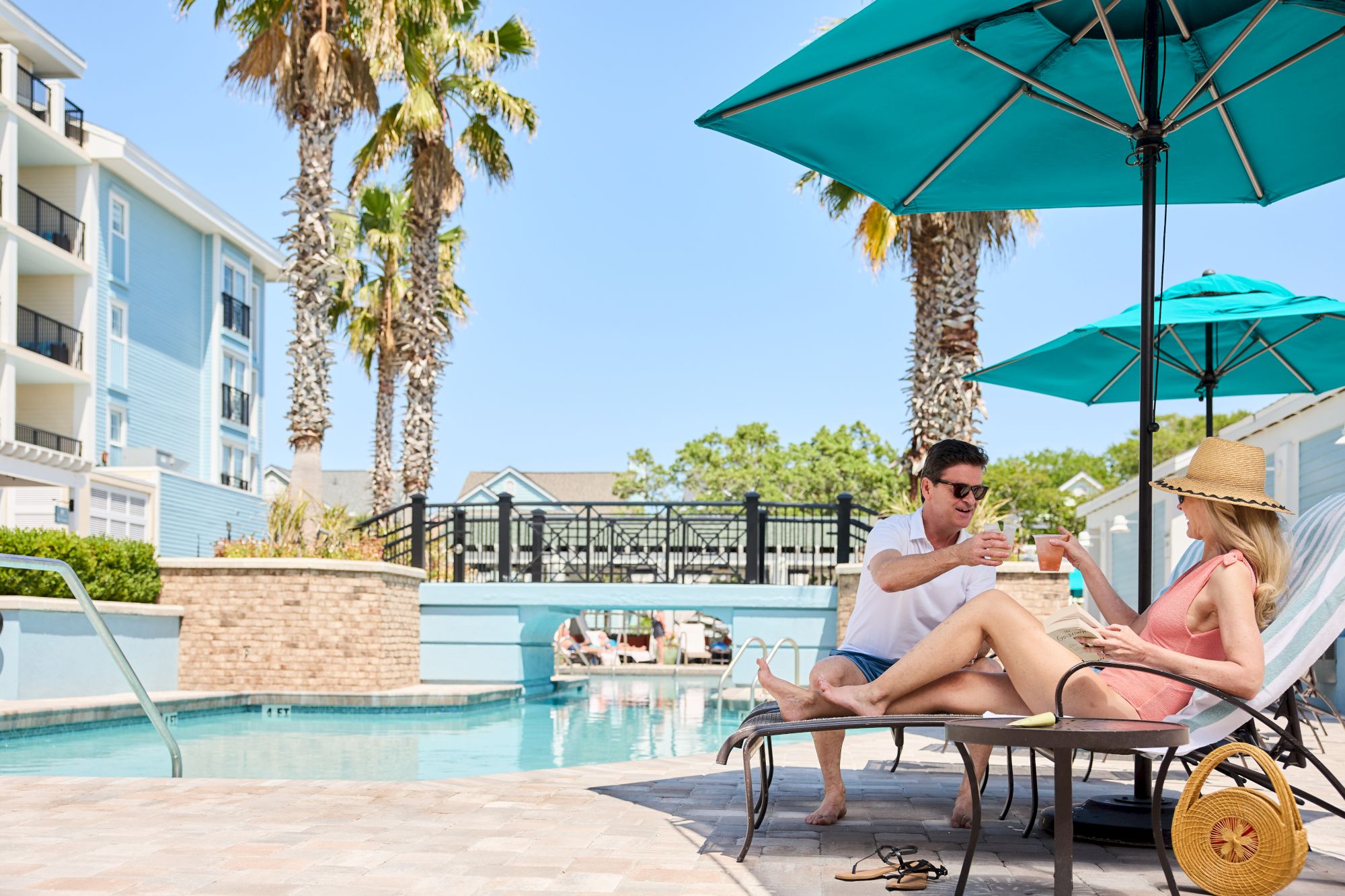 A couple lounges by a pool under umbrellas, with palm trees and buildings in the background, creating a relaxed vacation atmosphere.