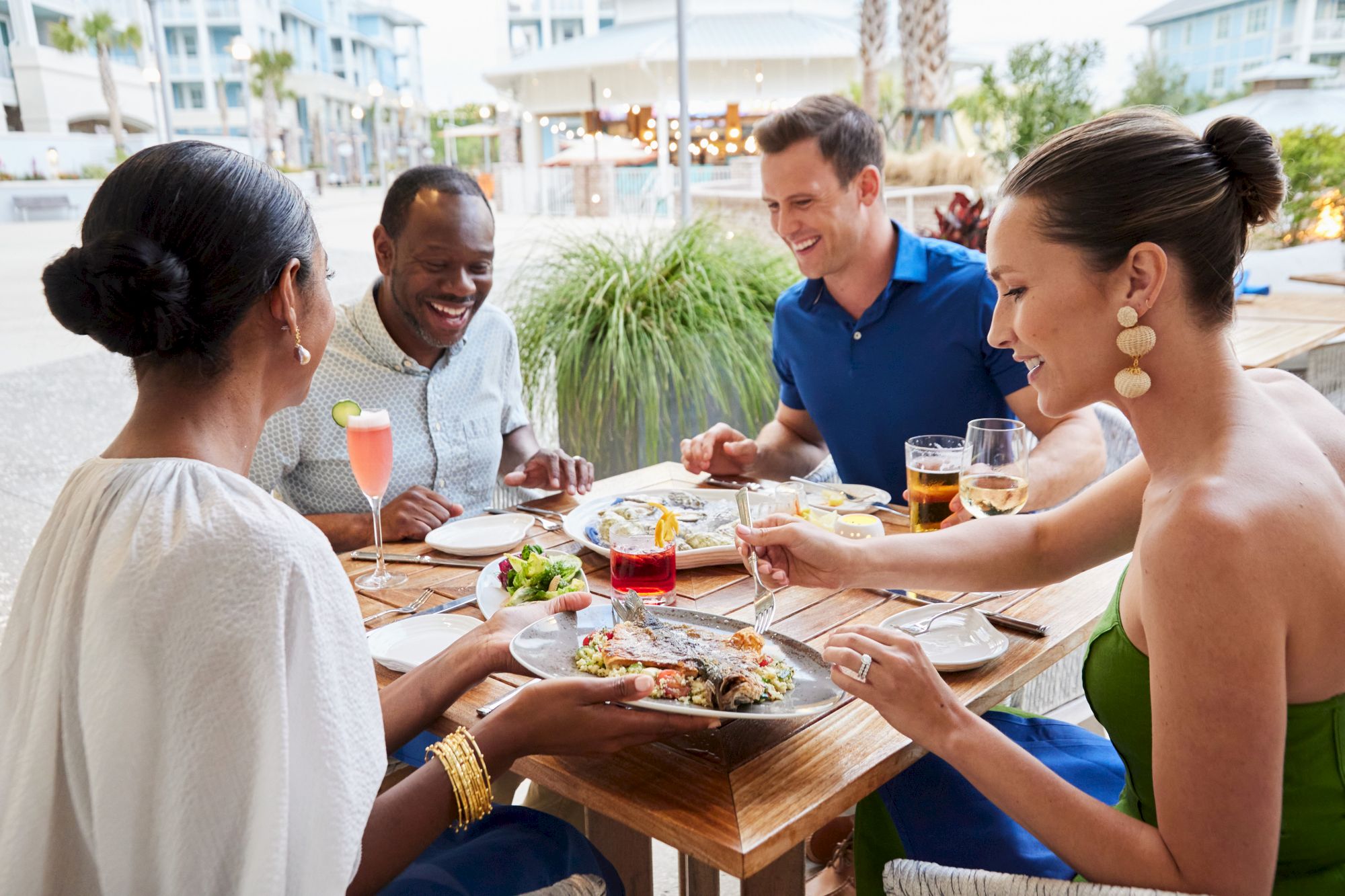 Four people are joyfully dining outdoors, enjoying drinks and food together at a wooden table.
