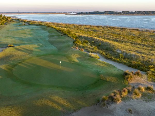 The image shows a golf course by the sea, featuring greens, sand bunkers, and a flagpole under a clear sky.