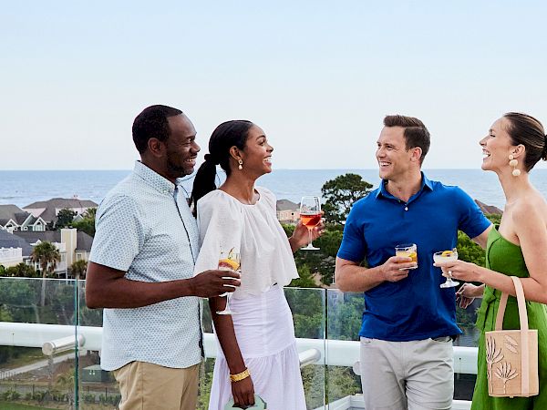 Four people are enjoying drinks and chatting on a rooftop with a scenic ocean view in the background.