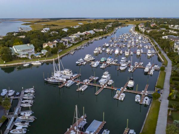 The image shows an aerial view of a marina with numerous boats docked along the waterway, surrounded by houses and greenery.