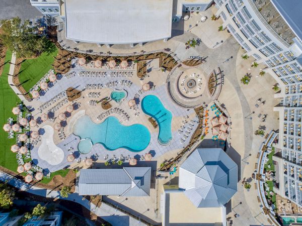 Aerial view of a resort pool area with various shaped pools, lounge chairs, umbrellas, and surrounding buildings in a sunny setting.