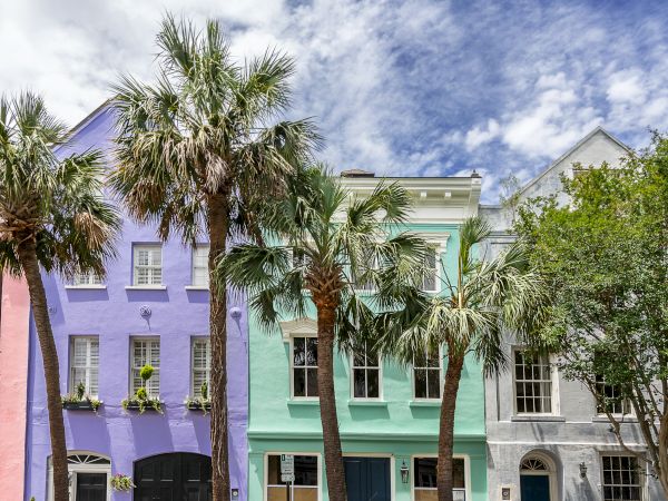 Colorful row houses in pastel shades of pink, purple, and green, lined with palm trees under a partly cloudy sky.
