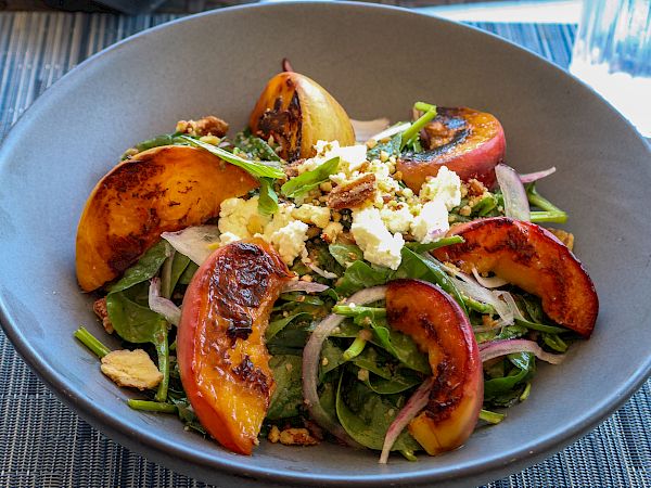 The image shows a grilled peach salad with greens, onions, and cheese, surrounded by plates of various appetizers on a blue table setting.