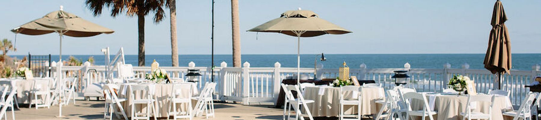 Outdoor event by the ocean with tables and chairs around a pool, decorated with palm trees and umbrellas.