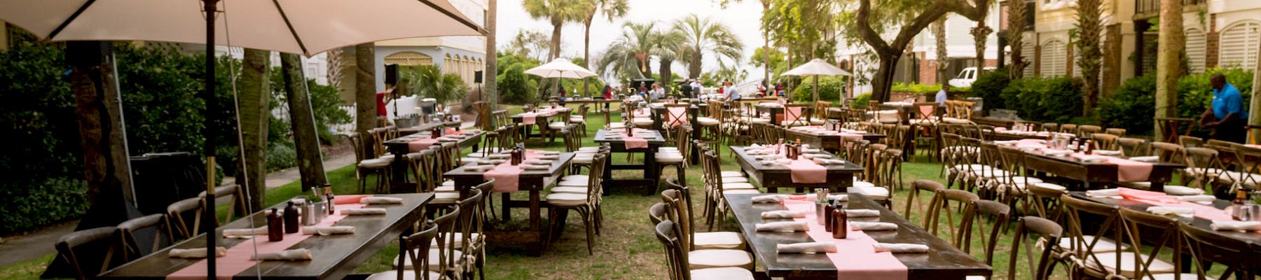 The image shows an outdoor dining setup with long tables, chairs, umbrellas, and pink table runners on a grassy area between buildings.