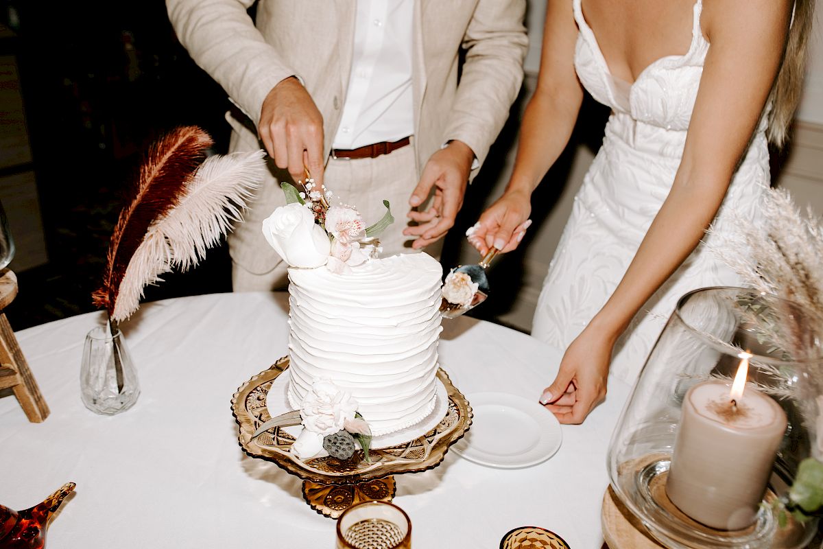 A couple is cutting a white tiered cake decorated with flowers. They are at a table with a candle and decorative feathers.