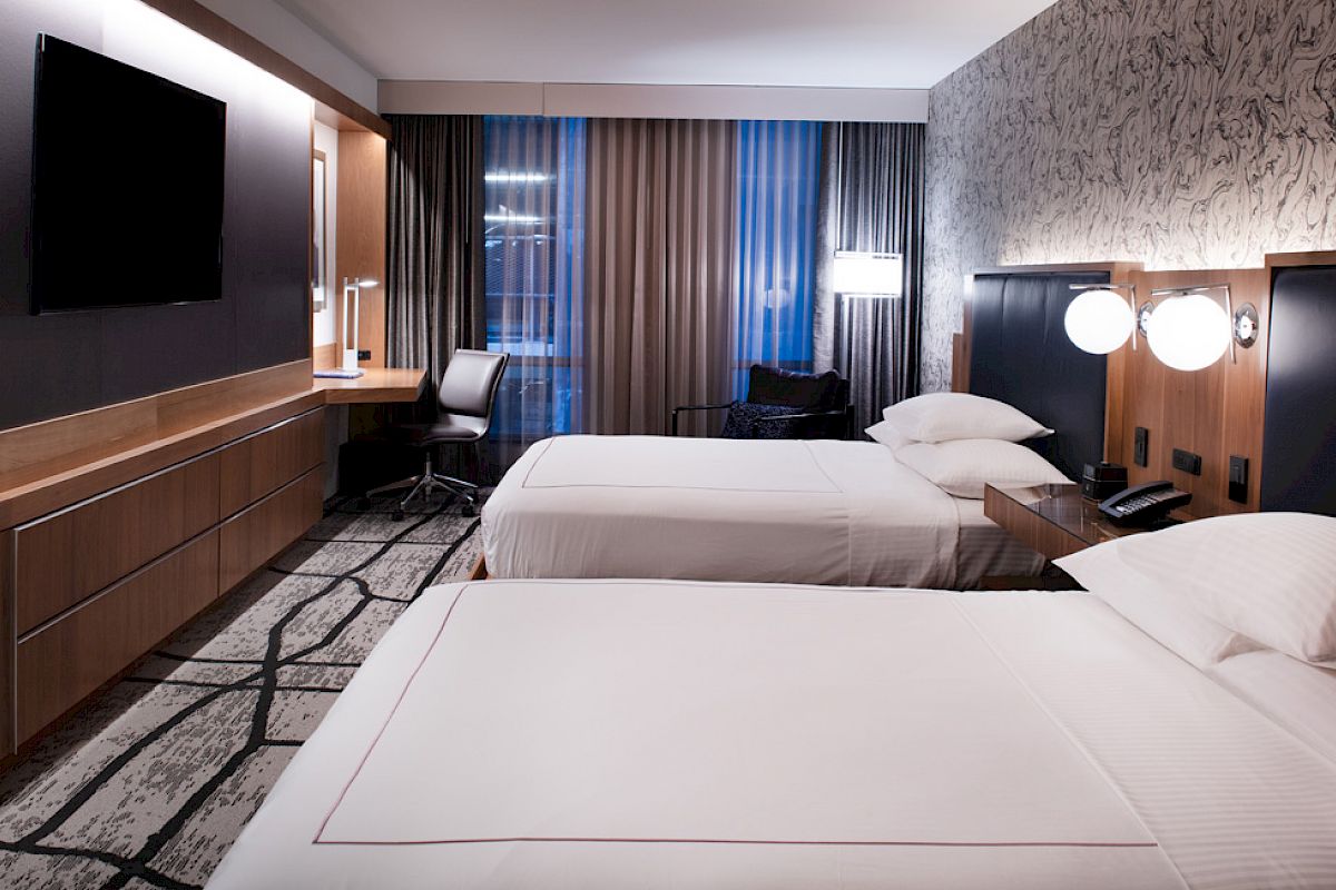 A modern hotel room with two beds, wall-mounted TV, desk, chair, and stylish lighting. The decor is sleek and contemporary.