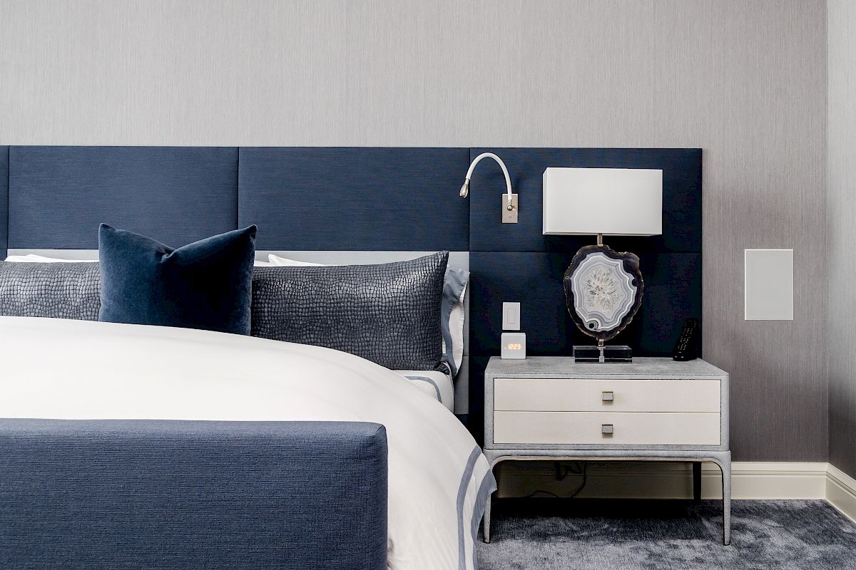 The image shows a modern bedroom with a blue headboard, white bed linens, a nightstand, lamp, and decorative pillow on a gray carpet.
