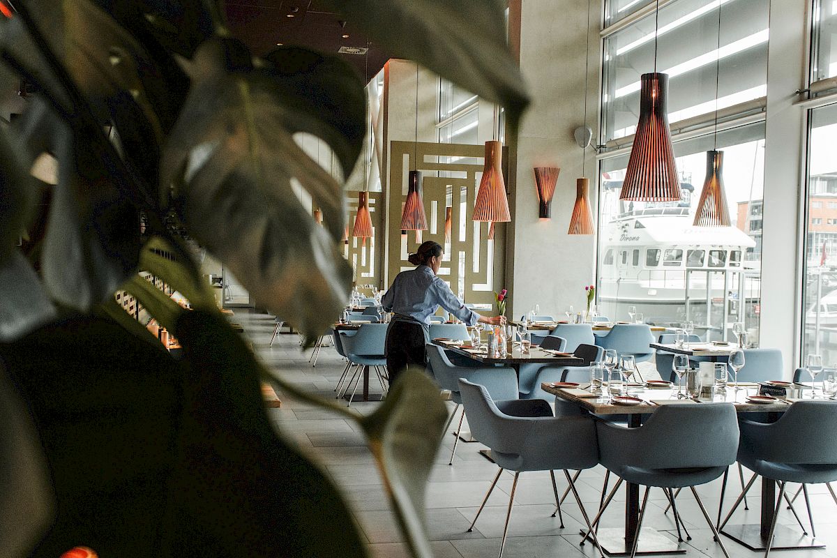 A person arranges a table in a modern restaurant with large windows and decorated lighting.