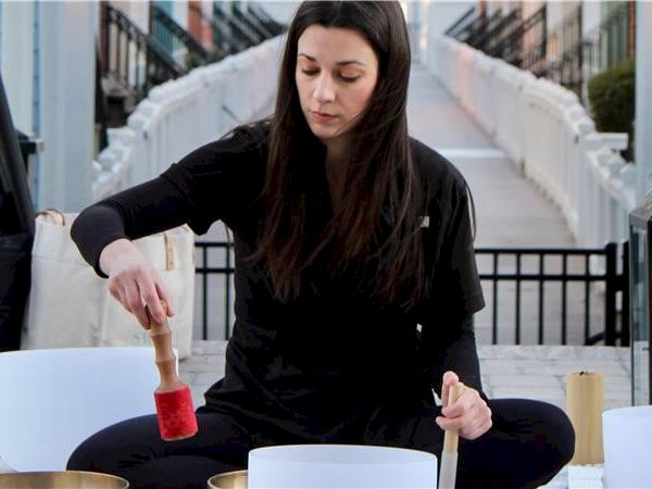 A person is playing singing bowls outdoors, seated, holding a mallet. The setup includes several bowls on the ground in front of them.