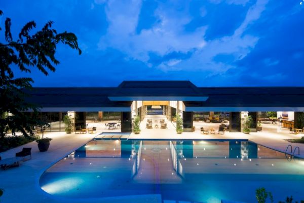 A modern building with an illuminated outdoor pool at night, surrounded by seating areas, under a deep blue sky.
