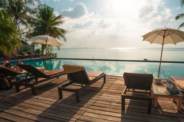 A serene poolside view with lounge chairs, umbrellas, and a tropical backdrop of palm trees and the ocean under a bright sky.