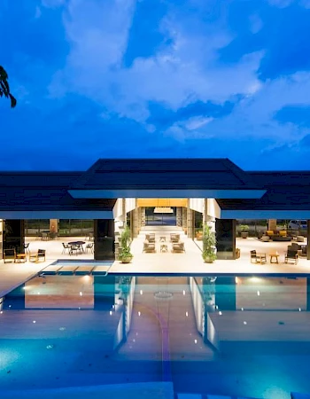 A modern building with large glass windows, lit warmly, is seen at night, overlooking an expansive swimming pool.