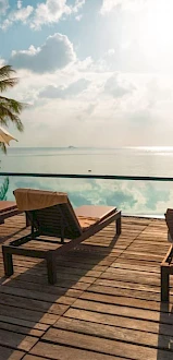 A serene poolside view with lounge chairs, umbrellas, palm trees, and a stunning horizon over calm waters under a partly cloudy sky.