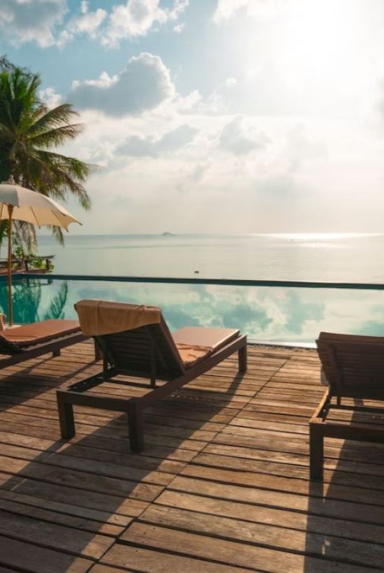 A serene beach scene with lounge chairs, umbrellas, and a pool overlooking the ocean, surrounded by palm trees under a sunny sky.