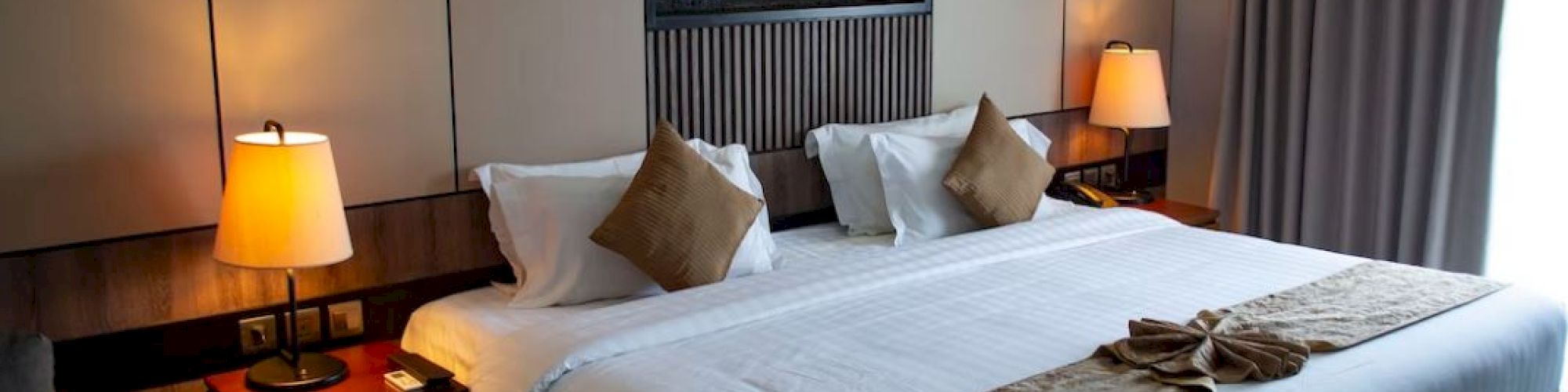 A neatly arranged hotel room features a large bed with brown and white linens, bedside tables with lamps, and a decorative wall panel.