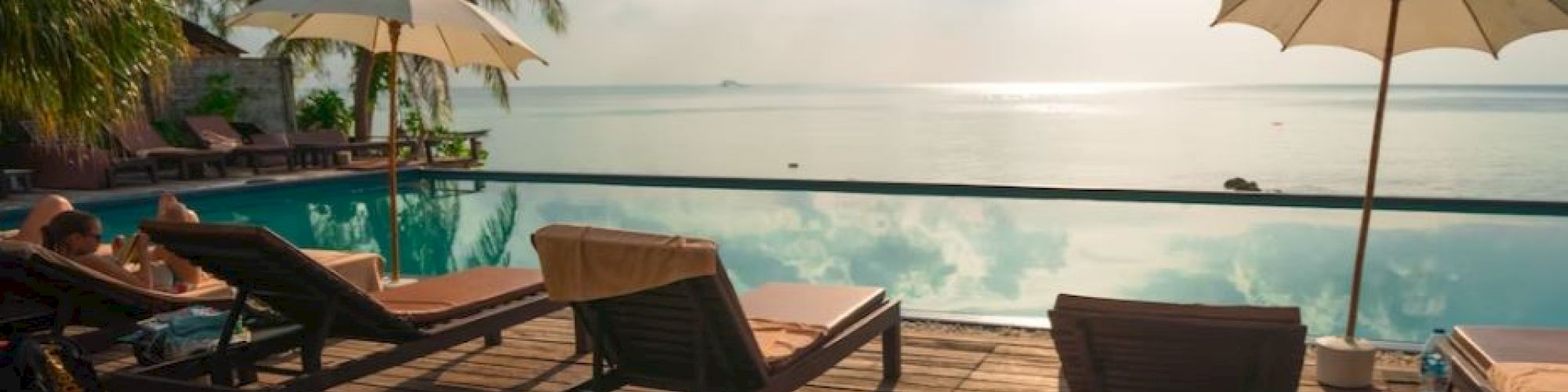 A tranquil poolside scene with lounge chairs, umbrellas, palm trees, and a serene view of the ocean under a partly cloudy sky.