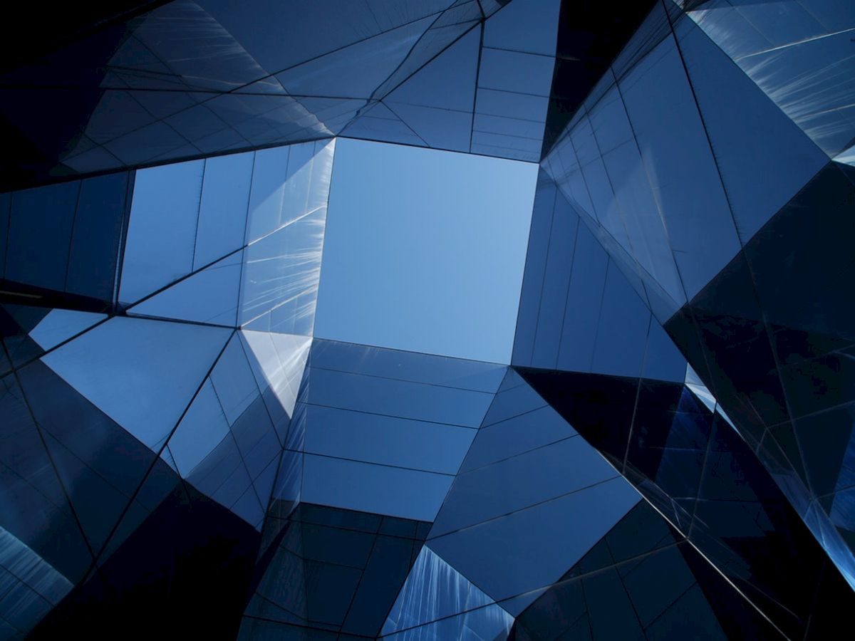 A view of a blue sky framed by mirrored, angular architectural structures, creating a geometric and abstract visual effect.