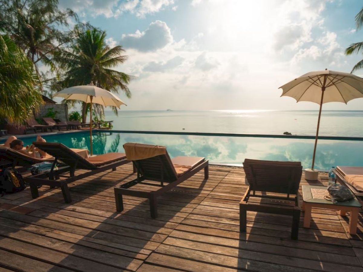 A serene poolside scene with lounge chairs, palm trees, umbrellas, and a view of the water under a partly cloudy sky.