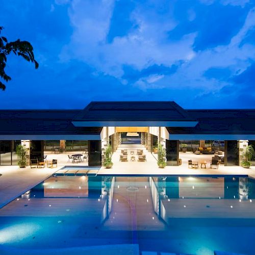 A luxurious building with an outdoor pool, lit up at night, surrounded by chairs and tables under a vibrant blue sky with clouds.