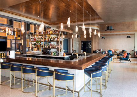 The image shows a stylish bar area with a wooden counter, blue barstools, and pendant lights. The shelves are stocked with various beverages.