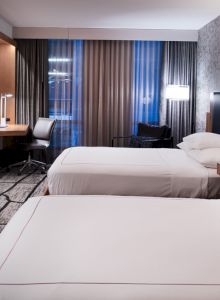 The image shows a modern hotel room with two beds, a flat-screen TV, a desk, and stylish decor including patterned wallpaper and curtains.