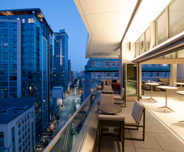 A modern cityscape at dusk features tall buildings and a stylish balcony with seating and tables overlooking the street below.