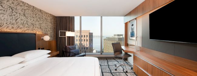 A modern hotel room features a large bed, desk, chair, wall-mounted TV, and a window with a city view, decorated with wood and patterns.